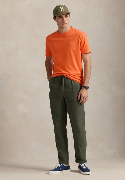 Fiatal férfi áll, narancssárga rövid ujjú pólót, olívazöld zsinóros nadrágot, kék sportcipőt, zöld sapkát és karórát visel.