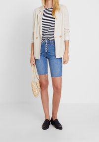 Beige liinapüks, mis on kantud triibulise must-valge pluusiga, koos siniste denim Bermuda lühikesed pükstega. Mustad sussid ja beež punutud kott.