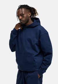 Navyblaues Hoodie aus weichem Stoff mit einer Fronttasche, Kapuze mit Kordelzug und gerippten Bündchen. Das Model steht mit der Hand am Kinn.
