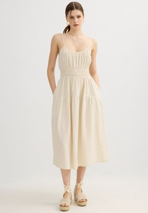 Joven con el pelo castaño recogido, que lleva un vestido midi sin mangas beige con cintura fruncida y sandalias cuña beige con lazos en el tobillo.