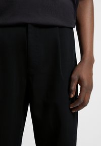 Pantalones plisados negros hechos de una tela suave, con una cinturilla ajustada y bolsillos, mostrados con una parte superior oscura y una mano descansando de lado.