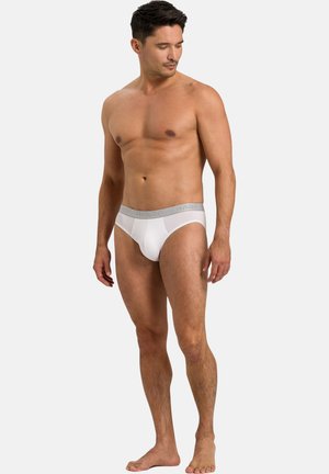 Uomo adulto in forma, in piedi a piedi nudi, indossa slip bianchi con una vita grigio chiaro, guarda in basso verso sinistra su uno sfondo semplice.