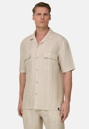 Mann trägt beiges Kurzarm-Hemd mit Brusttaschen und passende Hose, steht vor hellem, einfarbigem Hintergrund.