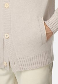 Ljusbeige stickad cardigan med stora runda knappar, ribbade muddar och en diskret fickdetalj, tillverkad av ett mjukt material för komfort.