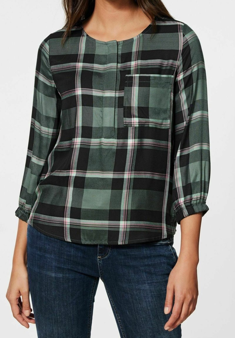 Chemise à carreaux vert, noir et blanc. Manches longues avec poignets élastiques, encolure boutonnée et poche poitrine. Texture de tissu lisse.