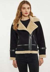Jachetă din blană shearing neagră cu un guler din blană sintetică bej, accente din piele, fermoare și curele. Are un design scurt cu mâneci lungi.