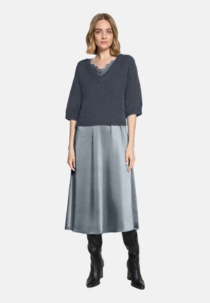 Frau trägt einen dunkelgrauen Strickpullover über einem silbernen Satin-Midirock und schwarzen Stiefeln mit Absatz, steht vor einem einfarbigen Hintergrund.