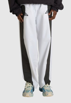 Persona che indossa pantaloni bianchi con pannelli laterali neri e scarpe da ginnastica blu e grigie, mani rilassate lungo i fianchi.