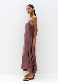 Robe maxi mauve avec bretelles spaghetti, ourlet en volants superposés, texture douce et silhouette fluide. Modèle portant des sandales plates.