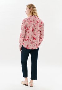 Donna in piedi con la schiena rivolta verso la macchina fotografica, che indossa una blusa a strisce floreali rossa e bianca, jeans scuri e décolleté beige.