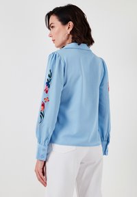 Light blue long-sleeve shirt with embroidered floral designs on sleeves, featuring a collar and buttoned cuffs. Soft fabric and relaxed fit.