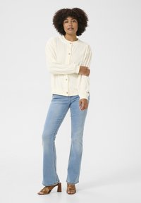 Cardigan crema con scollo rotondo e bottoni in legno, abbinato a jeans azzurri svasati e sandali con tacco marroni su uno sfondo neutro.