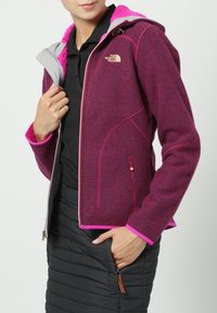 Burgundowa kurtka polarowa z kapturem, różowymi akcentami i fakturowanym materiałem. Posiada kieszenie zapinane na zamek oraz dopasowany krój.
