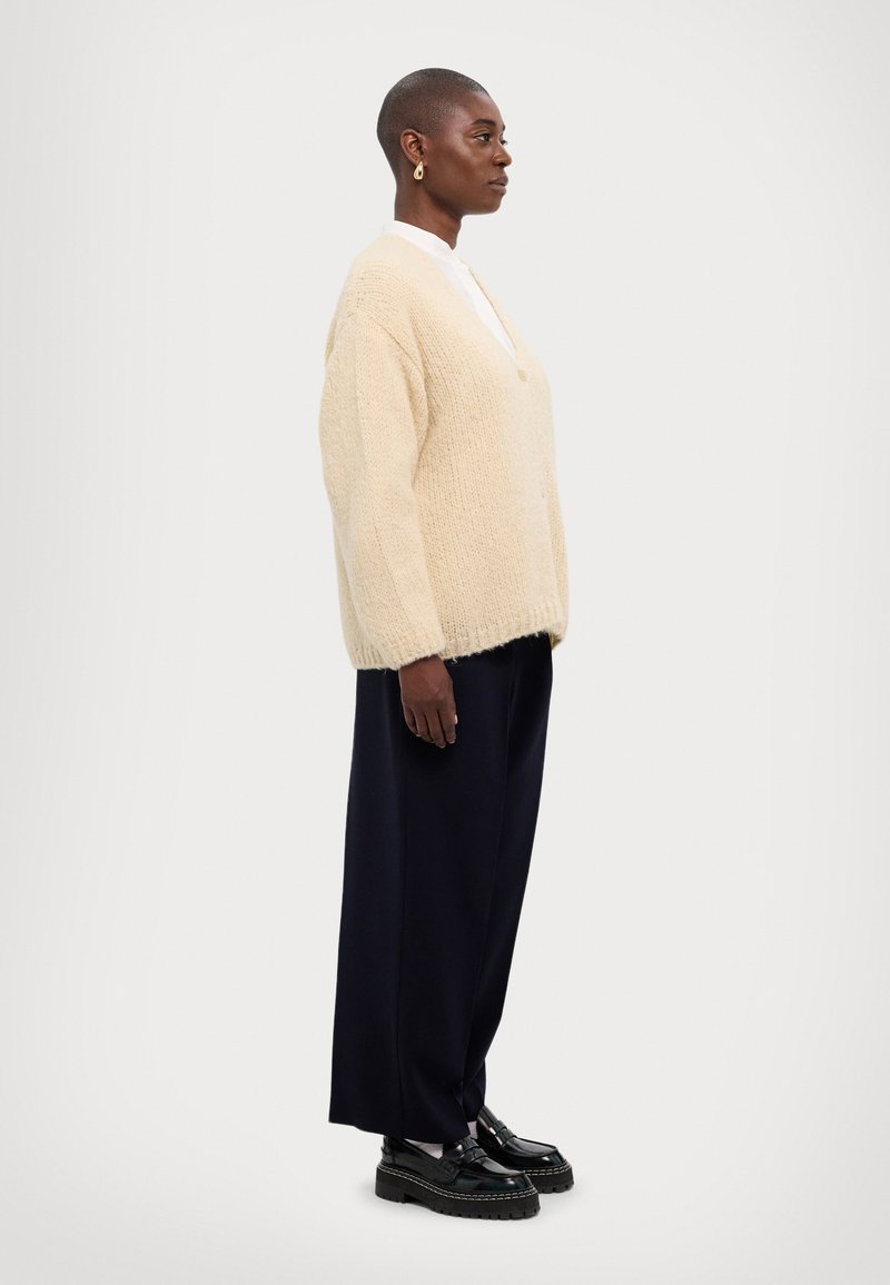 Cremfarbener Strickcardigan mit V-Ausschnitt, lockerer Passform und gerippten Bündchen, kombiniert mit dunklen, weit geschnittenen Hosen und klobigen schwarzen Slippers.