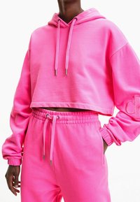 Rosa kort hoodie med snören och broderad detalj på ärmen, matchad med rosa joggers som har elastisk midja och fickor.