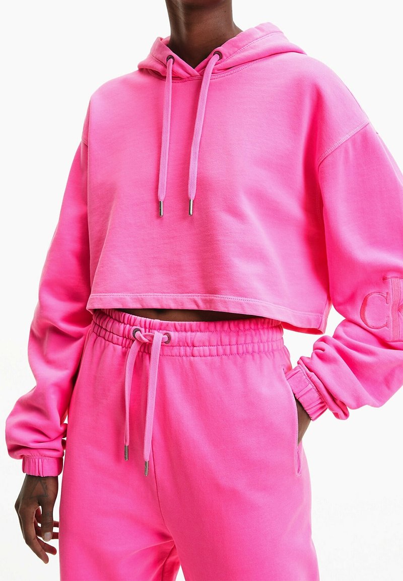 Rosa kort hoodie med snören och broderad detalj på ärmen, matchad med rosa joggers som har elastisk midja och fickor.