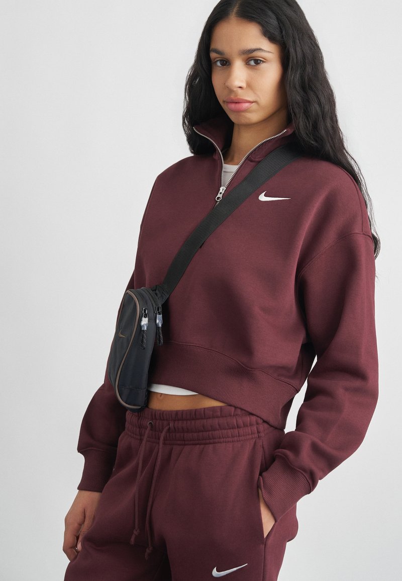 Giovane donna in tuta Nike color bordeaux con giacca zip-up e borsa a tracolla nera, in piedi davanti a uno sfondo chiaro e uniforme.