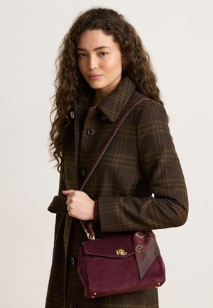 Femme aux longs cheveux bouclés portant un manteau à carreaux marron et tenant un sac à main bordeaux avec un logo doré et un foulard attaché.