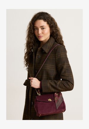 Femme aux longs cheveux bouclés portant un manteau à carreaux marron et tenant un sac à main bordeaux avec un logo doré et un foulard attaché.