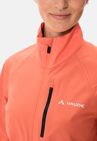 Korallfärgad väderjacka med hög krage, svart dragkedja och en ficka på vänster bröst. Har en vit logo på framsidan.