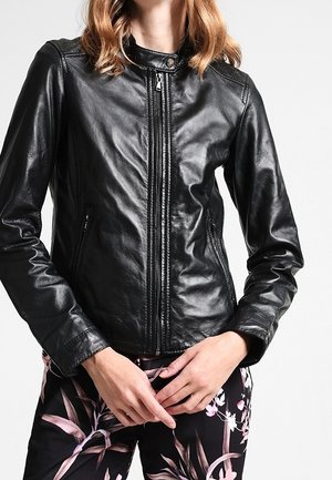Femme portant une veste en cuir noire ajustée avec fermeture éclair à l'avant et un pantalon noir fleuri, les mains jointes à la taille.