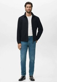 Chaqueta de navegación con cremallera y cuello extendido, combinada con jeans de mezclilla azul claro y zapatos de cuero negros. Diseño simple y limpio, sin patrones.
