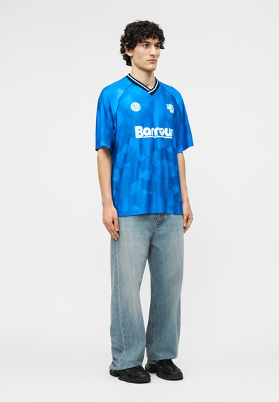 Άνδρας με σγουρά μαλλιά, που φοράει μπλε αθλητική φανέλα με μοτίβα και το λογότυπο "Barrow", ανοιχτόχρωμα ξεθωριασμένα τζιν και μαύρα αθλητικά παπούτσια, στέκεται μπροστά σε απλό φόντο.
