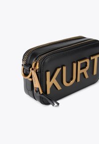 Sac en cuir noir avec ferrures dorées, présentant de grandes lettres embossées "KURT" sur le devant. Fermeture à double fermeture éclair et détail d'étiquette visible.