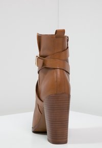 Hnědá kožená kotníková bota s vysokým hranatým podpatkem, páskem kolem kotníku se sponou a texturovaným designem dřevěného podpatku.