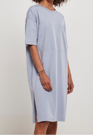 Femme portant une robe ample bleu clair à manches courtes, longueur genoux, fendue sur les côtés et avec un col rond, debout devant un fond uni.