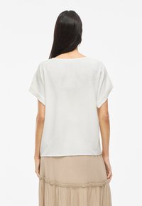 Top bianco a maniche corte con una vestibilità rilassata e una texture morbida. Il retro presenta un ampio scollo e viene abbinato a una gonna beige a strati.
