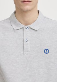 Polo shirt gris clair en tissu texturé avec un col, ornée de boutons bleus et d'un logo bleu brodé sur la poitrine.