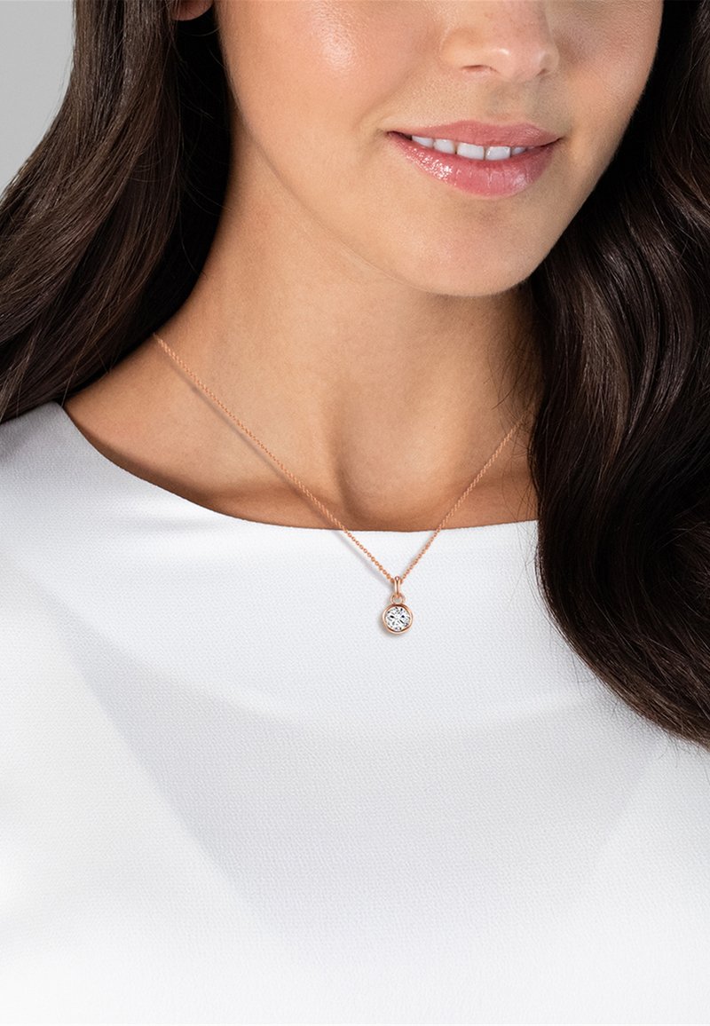 Woman wearing a rose gold necklace with a round clear gemstone pendant on a fine chain, dressed in a white top.