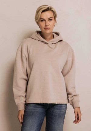 Hellbeiger Kapuzenpullover mit lockerem Schnitt, weicher Textur und gerippten Bündchen. Kombiniert mit blauen Jeans, vor einem neutralen Hintergrund präsentiert.