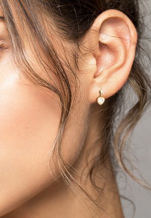 Goldfarbene, herzförmige Ohrringe mit einem klaren Edelstein, die von einem runden Stecker hängen und ein Nahaufnahme eines Ohrs sowie welliges Haar ergänzen.