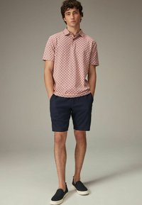 Tânăr îmbrăcat cu un tricou polo cu mânecă scurtă roz cu model, pantaloni scurți bleu marin și pantofi negri slip-on, stând cu mâinile în buzunare.