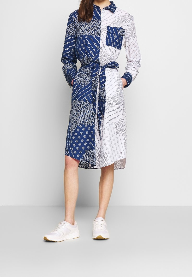 Robe chemise à motifs bleus et blancs avec un col à bouton, des manches longues, une ceinture assortie et des poches latérales. Associée à des baskets blanches.