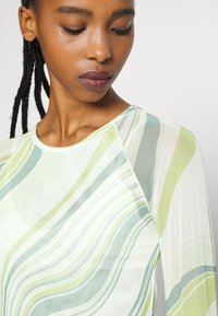 Blusa transparente de mangas longas com padrões ondulados em tons de verde claro e bege suave. Decote redondo e textura suave, apresentando variações de cor subtis.