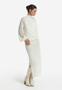 Top bianco a maniche lunghe con collo alto e maniche trasparenti, abbinato a una gonna lunga aderente con spacchi laterali. Completo con stivali beige al ginocchio.