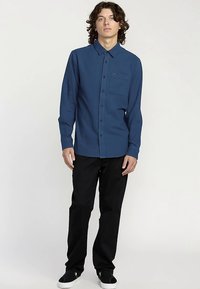 Camicia blu navy con bottoni, colletto e taschino sul petto, abbinata a pantaloni neri e sneakers nere. Tessuto liscio, maniche lunghe.