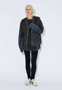 Tmavě modrá denimová vesta s dvěma předními kapsami, nošená přes černé tričko s dlouhým rukávem. Doplněno o černé legíny a bílé tenisky.