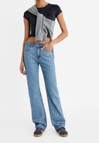 Top corto negro de manga corta combinado con jeans de pierna ancha de color azul claro, complementado con un suéter gris atado en la cintura. Un calzado casual completa el look.