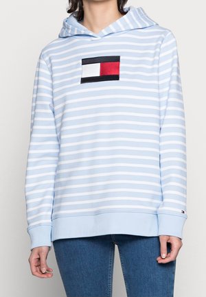 Osoba ubrana w bluzę z kapturem w jasnoniebieskie i białe poziome paski z czerwoną, białą i czarną prostokątną naszywką na piersi, w połączeniu z niebieskimi dżinsami.