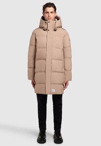 Abrigo acolchado color beige con capucha, que cuenta con cierre de cremallera y botones a presión. Textura acolchada gruesa, bolsillos laterales y una pequeña etiqueta de la marca en el dobladillo.