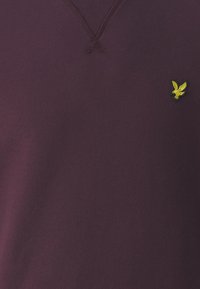 Jersey de cuello en V hecho de tela burdeos, con un cuello acanalado y un pequeño emblema de pájaro verde y amarillo en el pecho.