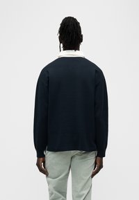 Μπλε πουλόβερ πλεκτό σε σκούρο μπλε χρώμα με στρογγυλή λαιμόκοψη, χαλαρή εφαρμογή και μακριά μανίκια, συνδυασμένο με ανοιχτό πράσινο παντελόνι με υφή και λευκό πουκάμισο με γιακά από κάτω.