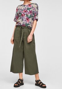 Top floreale con maniche corte, caratterizzato da tonalità viola e rosa, abbinato a pantaloni larghi in lino verde e sandali neri con cinturini.