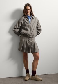 Chaqueta bomber gris con ribetes blancos, combinada con una falda plisada gris. Camisa azul clara debajo, con zapatillas beige y calcetines burdeos.