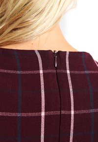 Tela burdeos con un patrón de cuadros; cuenta con un cierre de cremallera en la parte trasera y una textura suave. Acentuada con finas líneas blancas y azul marino.