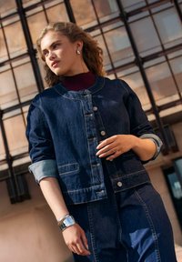 Veste en denim foncé cintrée avec un col rond, des boutons argentés et des manches retroussées, associée à un pull col roulé bordeaux et un bracelet argenté.